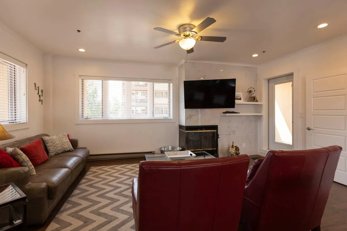 A cozy living room with a comfortable sofa, red accent chairs, a coffee table, and a large television. Windows provide natural light, creating a warm and inviting space for relaxing or socializing after a productive day. The layout encourages downtime—whether watching TV, reading, or hosting an informal meeting.