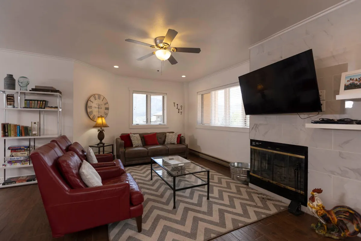 The image showcases a warm and inviting living area featuring bold red leather sofas arranged around a glass coffee table atop a stylish zigzag patterned rug. The modern fireplace glows at one end, promising cozy evenings, while a sleek television offers entertainment. The space exudes comfort and sociability, perfect for relaxing, reading, or gathering with loved ones after a day in the Colorado mountains.