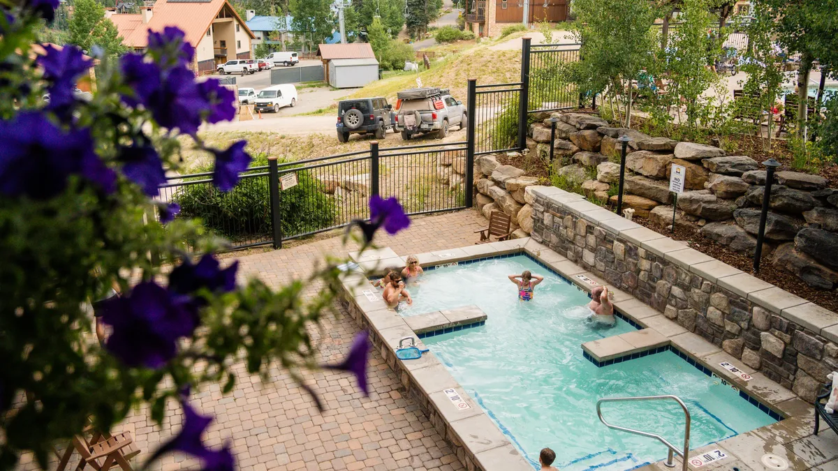 A cheerful outdoor swimming pool scene, where children and families enjoy swimming surrounded by flowers and a lush mountain landscape. The vibrant atmosphere highlights relaxation and socializing after a day of adventure.