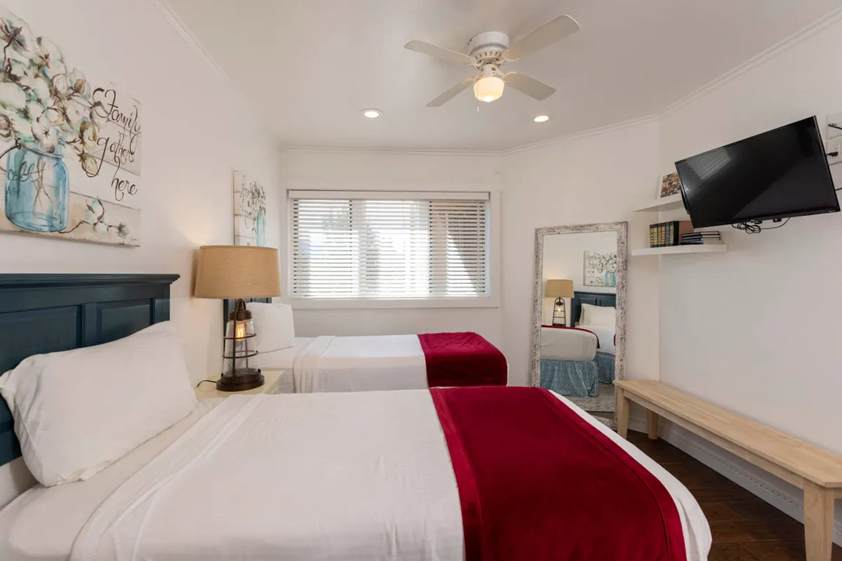 A comfortable bedroom with two beds adorned with red throws, decorative wall art, a table lamp for soft lighting, and a television, offering a flexible and welcoming setup for families or friends.