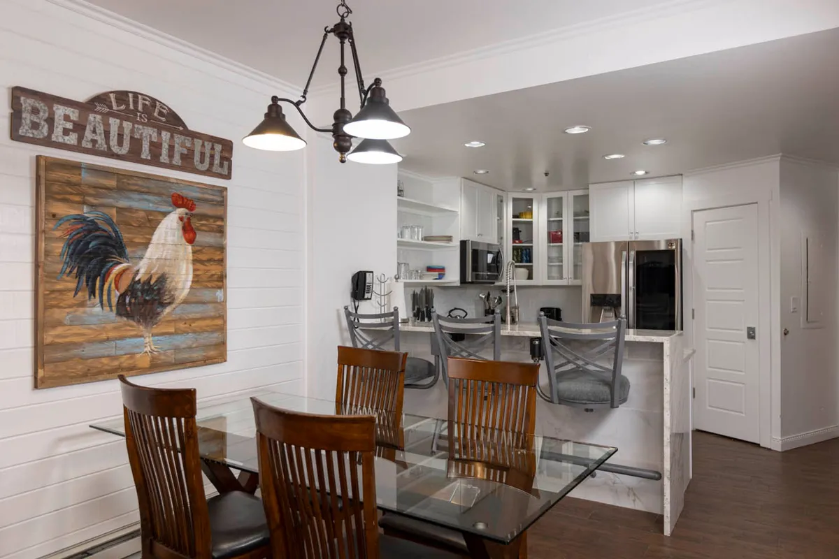 This image captures the open dining area with a prominent rooster artwork above the dining table, bar stools at the kitchen counter, and glimpses of kitchen appliances. The setup encourages social gatherings, family meals, and offers visual continuity between dining and kitchen spaces.