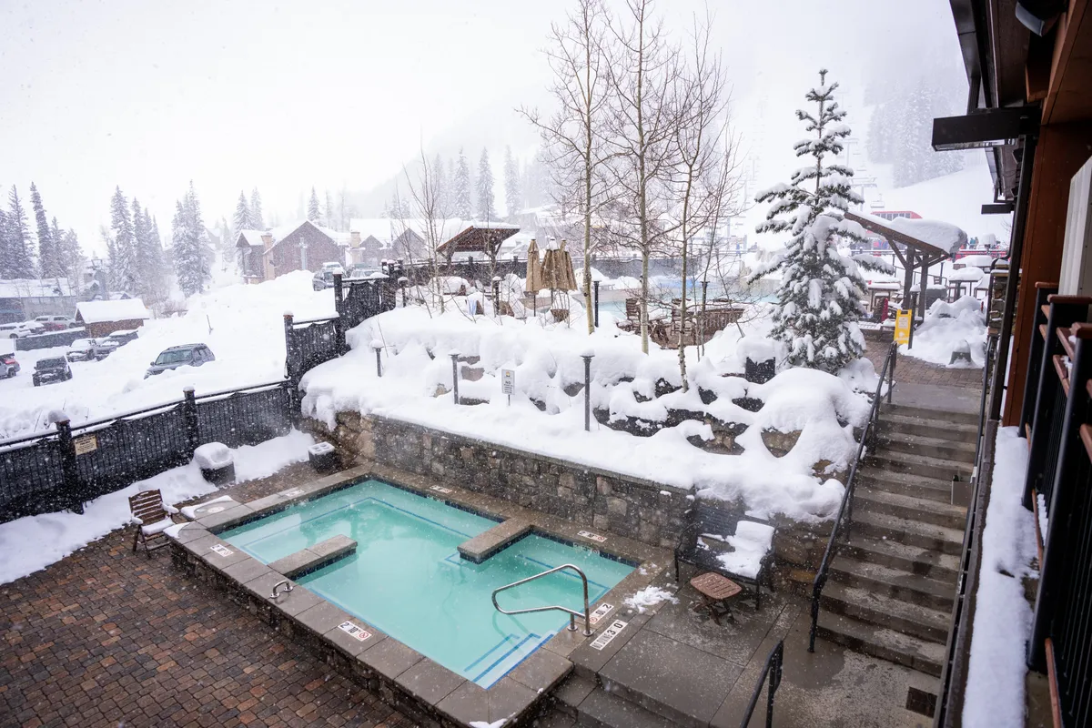 This image displays an inviting hot tub set amidst a snowy pine landscape, with steps leading up to the steaming water. The setting is serene and luxurious, perfect for relaxing and taking in mountain views, especially as dusk descends.