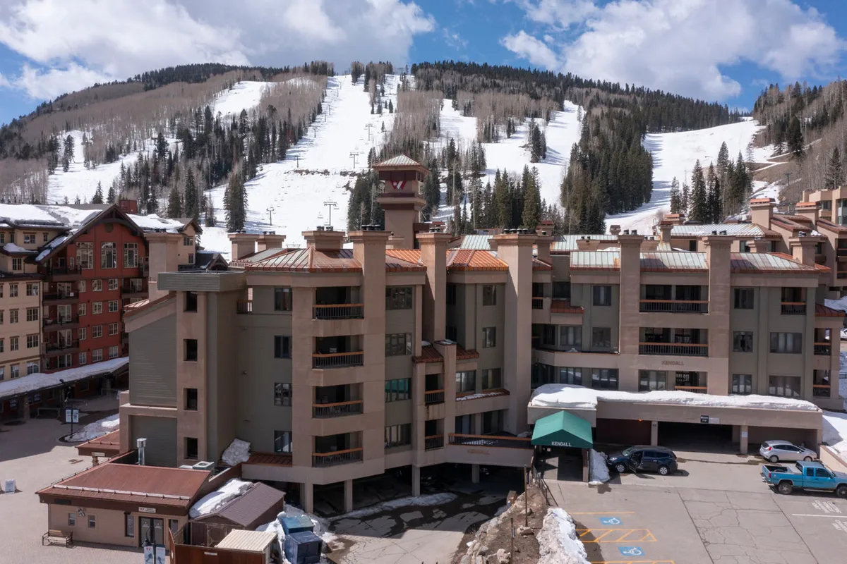This exterior image captures the building architecture with a view of the ski slopes, mountain backdrop, and visible parking area. The scene is lively and welcoming, conveying the proximity to both adventure and practical amenities.