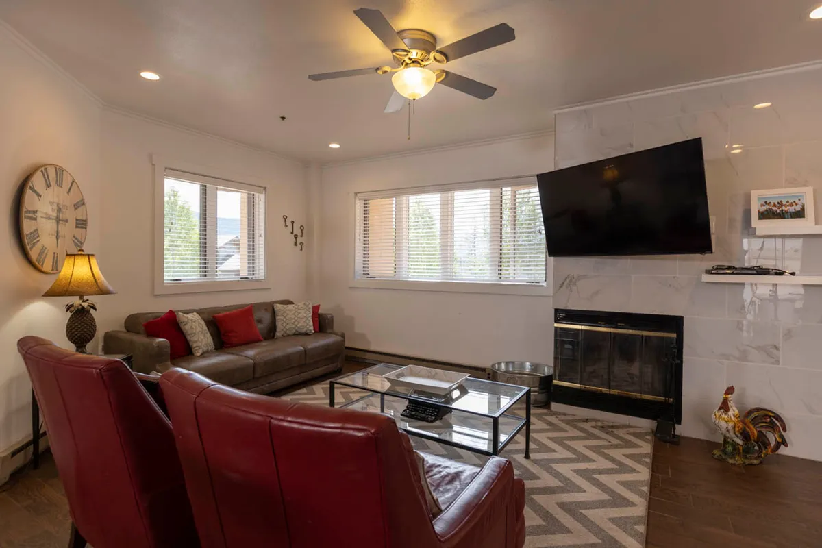 This interior image highlights a spacious living area featuring a plush sofa, red accent chairs, a central coffee table, and an inviting wood-burning fireplace. The television and relaxed seating arrangement create a warm, communal atmosphere ideal for cozy evenings, group conversations, or relaxing after outdoor adventures.