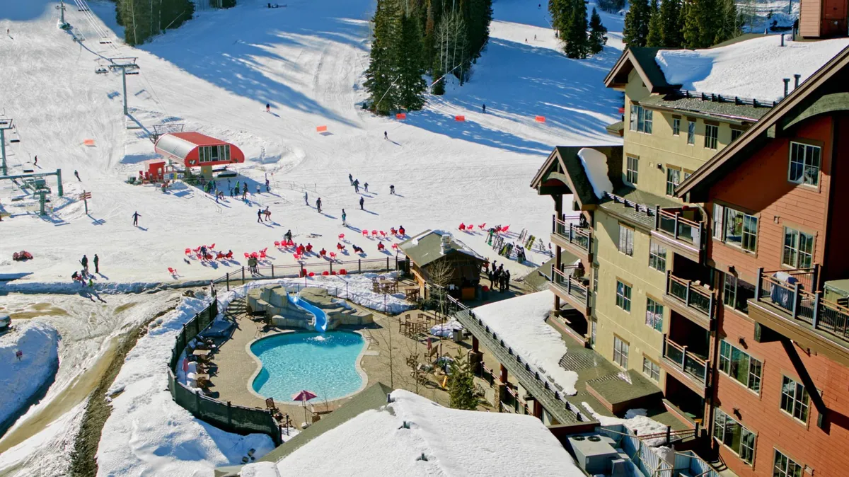 The image features an outdoor heated pool adjacent to a lodge building, with ski slopes and a ski lift visible in the background. The pool area is lively, blending resort activity with mountain scenery.