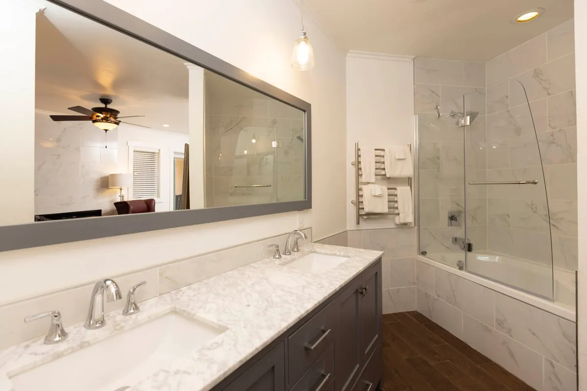 A modern bathroom featuring a double sink vanity with a large mirror, glass shower enclosure, marble countertop, and wood flooring, bathed in natural light for a refreshing start or end to the day.