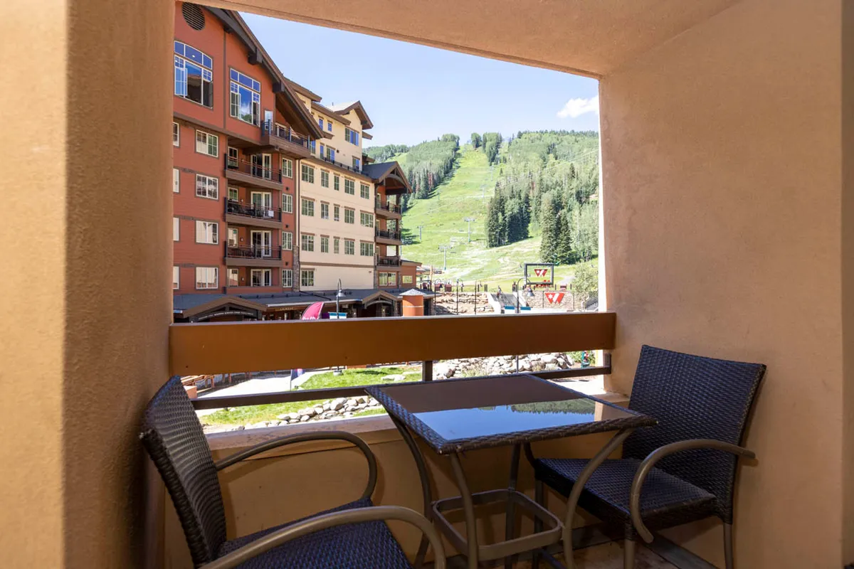 The image shows a balcony with comfortable outdoor seating, surrounded by buildings and offering a scenic mountain view. The lively village atmosphere is palpable, ideal for socializing or taking in the surroundings.