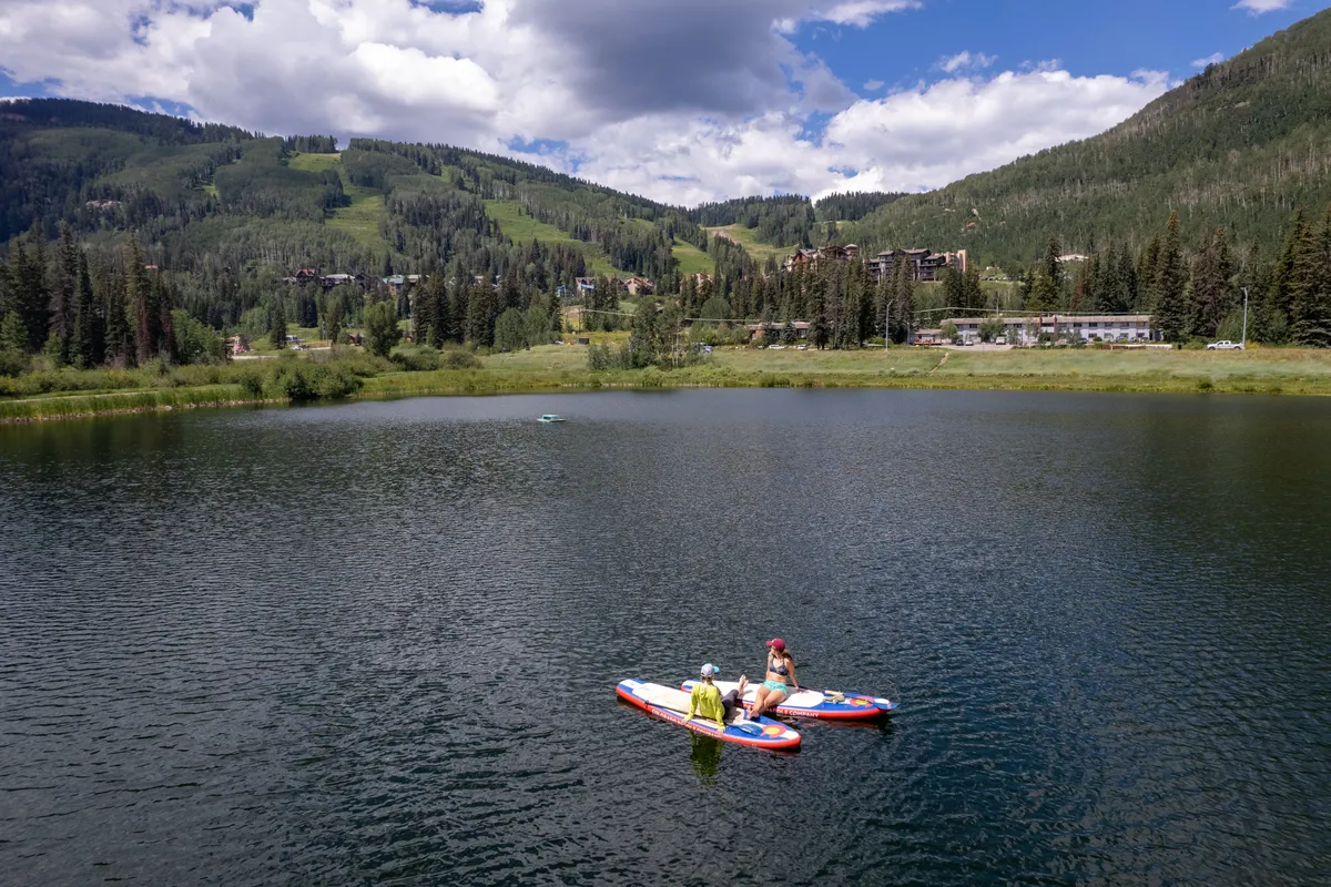 A lively scene featuring two people paddleboarding on a calm lake, surrounded by mountains. The playful mood and natural setting evoke fun, exploration, and family bonding through outdoor activities.